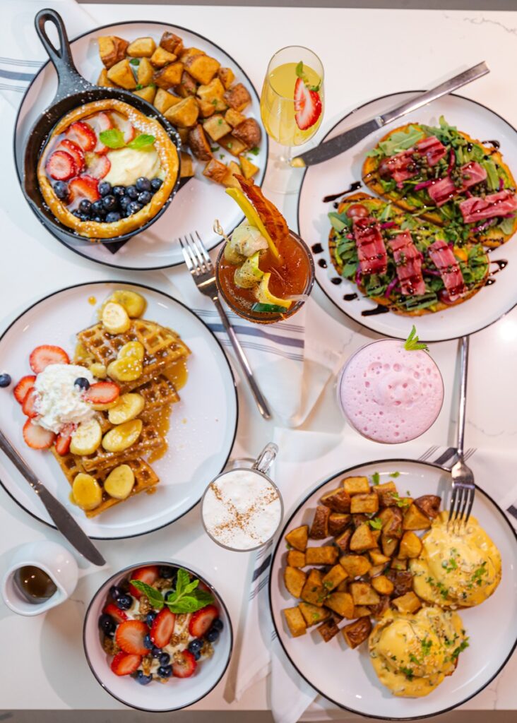 A table laden with various brunch dishes, including a Dutch baby pancake with berries and cream, waffles with bananas and strawberries, eggs Benedict with roasted potatoes, avocado toast with bacon, a parfait, and several cocktails.