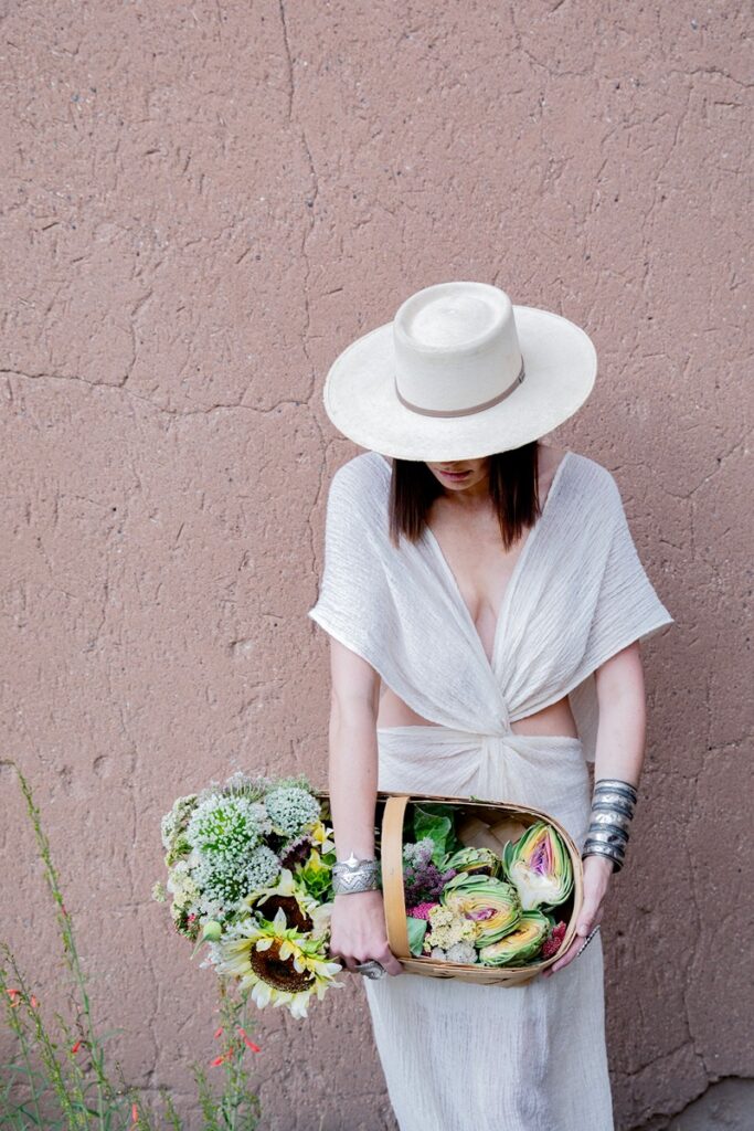 A person in a white hat and flowing white attire holds a basket overflowing with fresh produce, including various shades of green and purple cabbages, cut artichokes, and leafy greens, against a textured adobe-colored wall.
