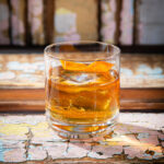 A short, clear glass holds a light brown clear the fog cocktail with ice and an orange peel twist. The glass is placed on a rustic wooden surface with peeling paint.