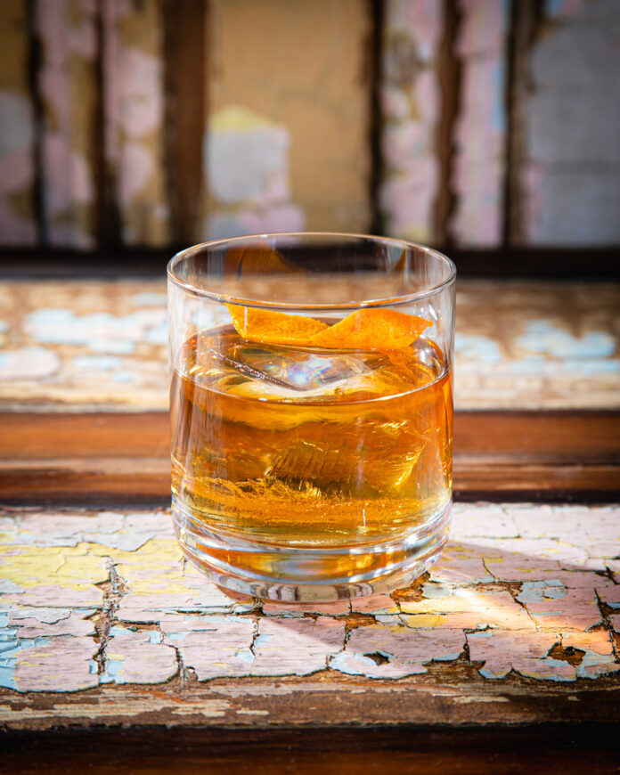 A short, clear glass holds a light brown clear the fog cocktail with ice and an orange peel twist. The glass is placed on a rustic wooden surface with peeling paint.
