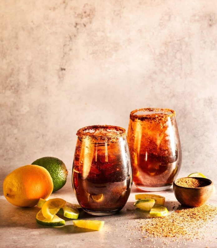Two Dark and Handsome Rum Moscow Mules sit in front of a painted brown background with Tajin rims and a lime and orange sitting next to the glasses.