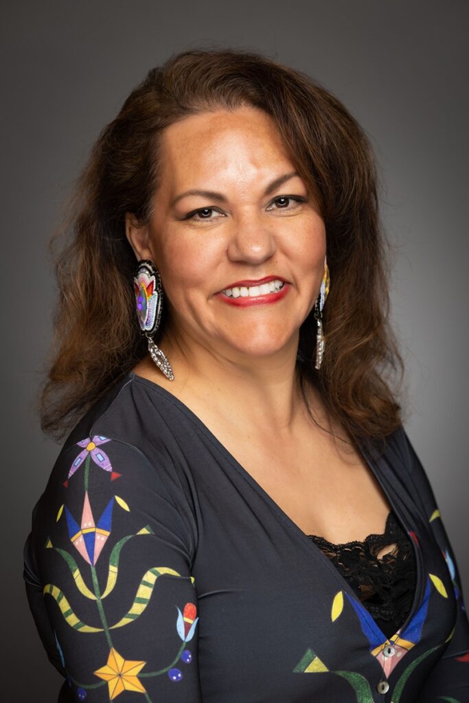 Dawn Houle with long, wavy brown hair, wearing a dark top with colorful embroidered patterns on the sleeves and large, ornate beaded earrings.