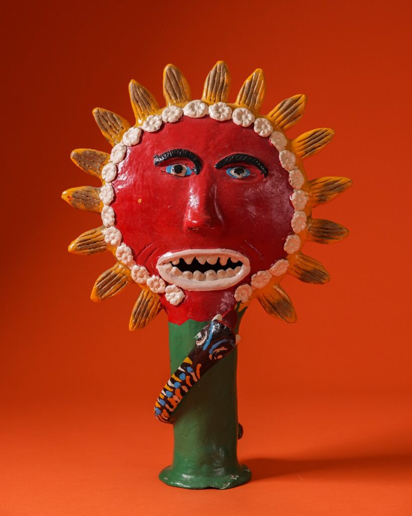An earthenware sculpture depicts a personified sun with a snake climbing towards it, featuring red, orange, and blue hues on a textured background.
