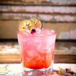 A vibrant pink cocktail is presented in a rocks glass filled with ice, garnished with two dried lime wheels and a small rosebud.