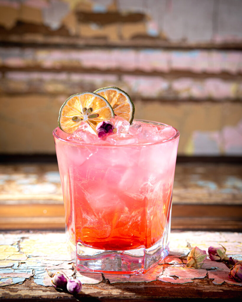 A vibrant pink cocktail is presented in a rocks glass filled with ice, garnished with two dried lime wheels and a small rosebud.