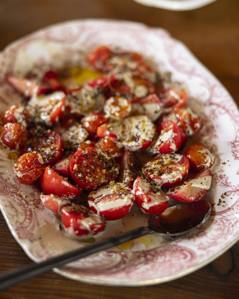 A bowl of vibrant red tomatoes, sliced and whole, is mixed with herbs and topped with a creamy tahini dressing and a sprinkle of dukkah.