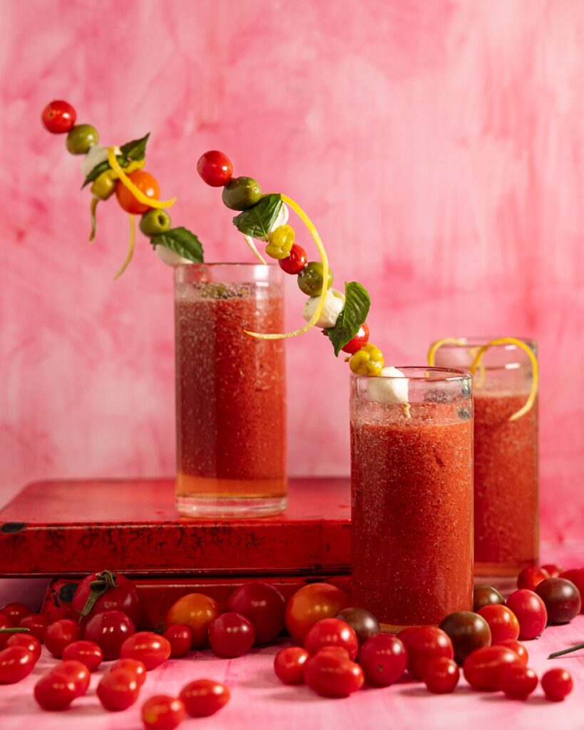 Three tall glasses filled with a vibrant red tomato-based drink, likely Bloody Marys, each garnished with skewers featuring colorful cherry tomatoes, mozzarella balls, green olives, basil leaves, and lemon zest spirals.