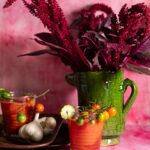 Two glasses of red gazpacho are on a pink surface, with cherry tomatoes and a garlic bulb on a wooden plate next to one glass.