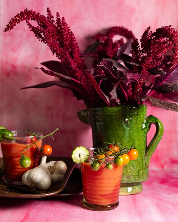 Two glasses of red gazpacho are on a pink surface, with cherry tomatoes and a garlic bulb on a wooden plate next to one glass.
