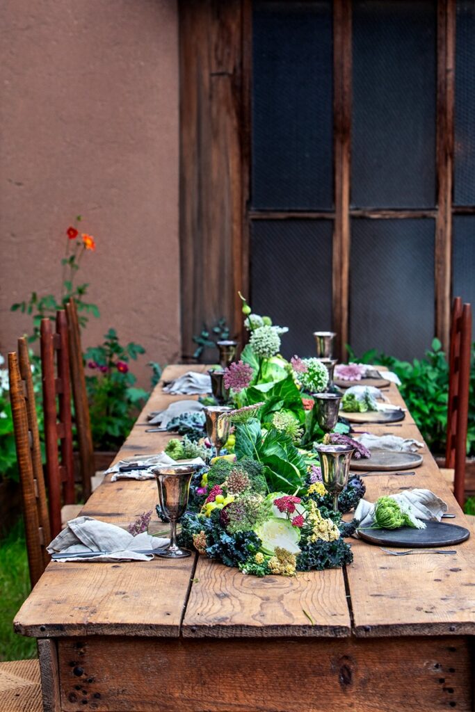 An outdoor dining table is laden with a vibrant edible tablescape featuring gifts from the garden including a variety of fresh produce like cabbages, kale, and other greens, along with silver goblets and dark plates set for a meal.