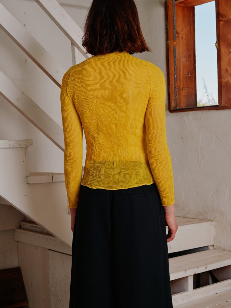 A person from the back, wearing a sheer, golden yellow Issey Miyake "twisted chiffon" shirt and black Tibi pants, standing near white stairs and a wooden-framed window.