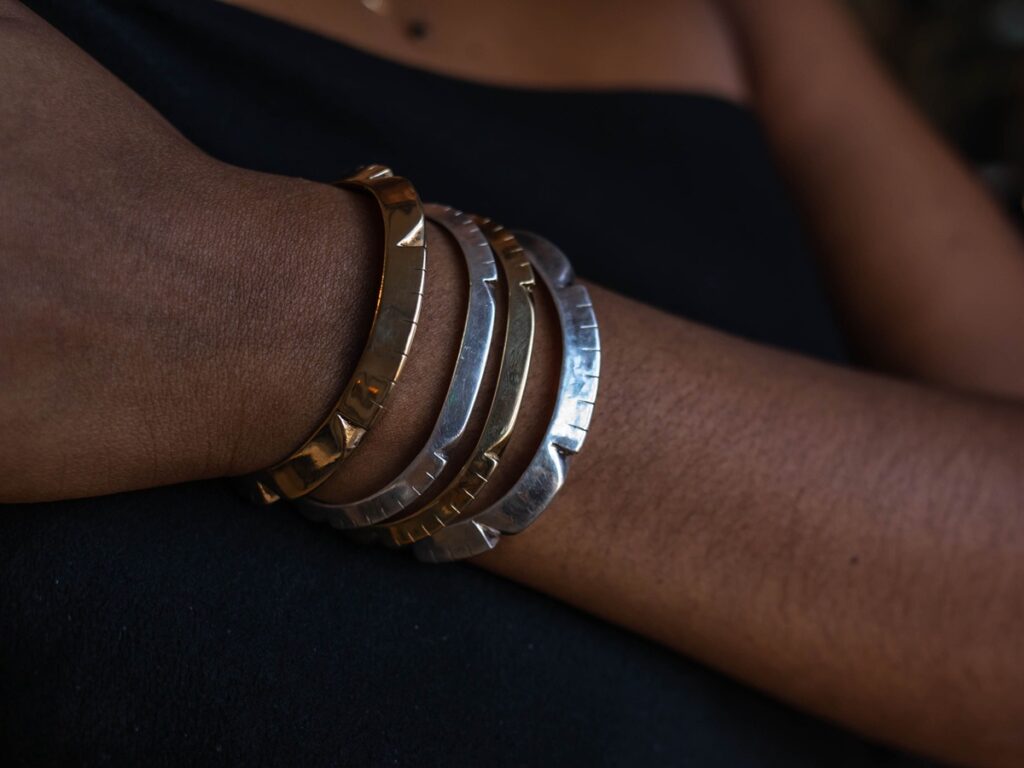 A close-up of a person's arm with darker skin wearing multiple silver and gold cuff bracelets with carved edges.