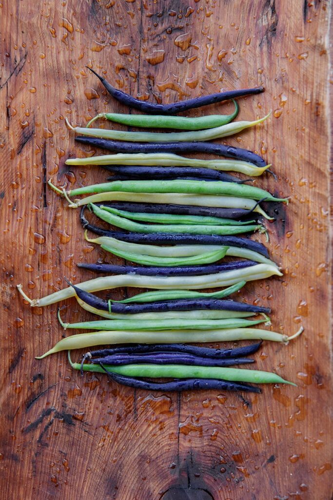 Assorted green, yellow, and purple string beans are laid out in rows on a damp wooden surface, showing water droplets.