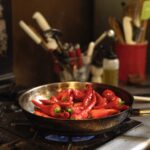A harvest mix of peppers gets cooked in a pan on a gas stove with utensils sitting in the background.