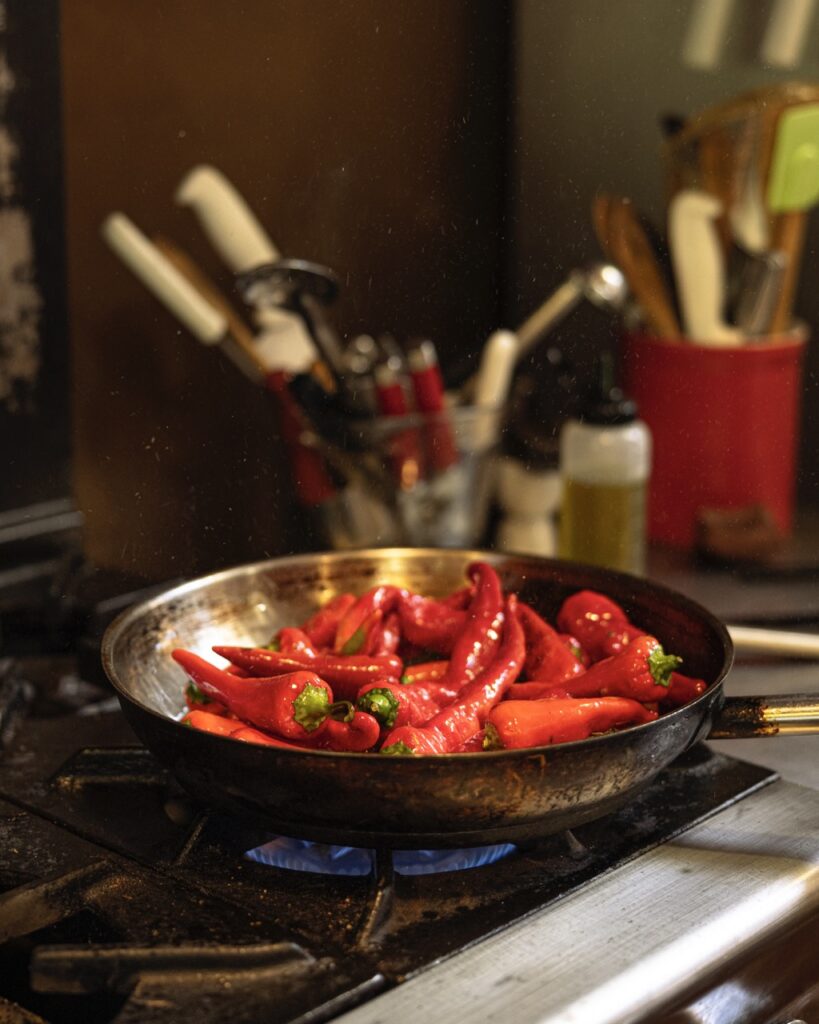 A harvest mix of peppers gets cooked in a pan on a gas stove with utensils sitting in the background.