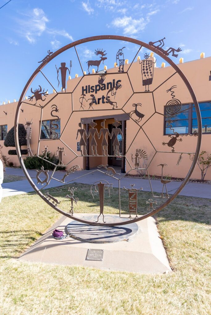 An abstract metal sculpture, possibly of Hispanic art, stands in front of a light brown building with the words "Hispanic Arts" on its facade.