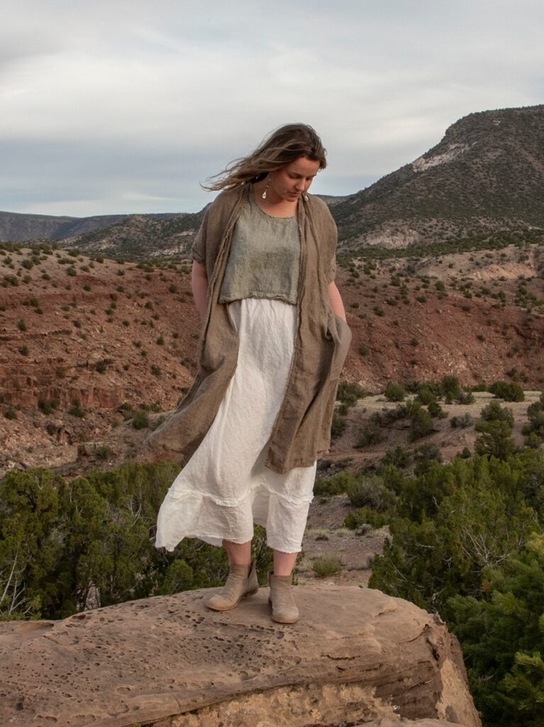 A woman with long brown hair stands on a rocky outcrop, wearing a light green linen crop top, a long white linen skirt, and a long brown linen wrap, with a desert landscape in the background.