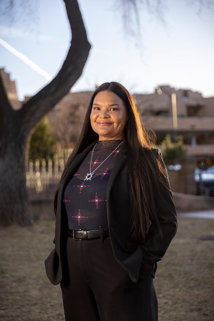 Jamie Schulze is pictured in front of Santa Fe Indian Market smiling outdoors. She is wearing a dark blazer over a dark top with a light-colored, geometric pattern and a necklace with a prominent, angular pendant.