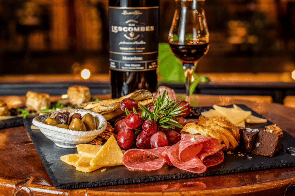 A bottle of Lescombes wine and a glass of red wine are displayed with a charcuterie board featuring cheeses, meats, olives, grapes, and crackers.