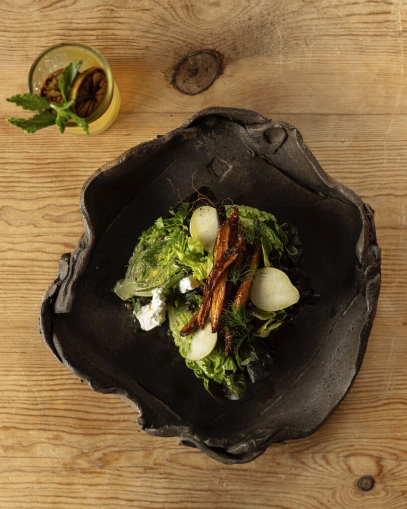 A black abstract bowl holds a little gem salad with roasted carrots and pear slices on top.