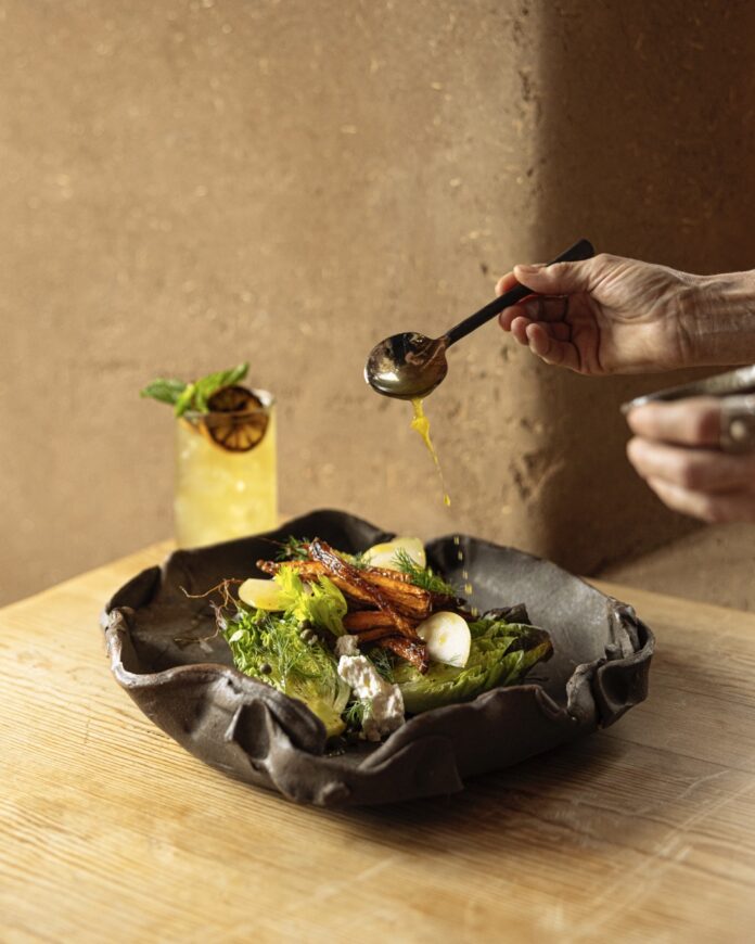 Little Gem Salad A person drizzles a spoonful of vinaigrette onto a little gem salad in a black abstract bowl with a yellow cocktail sitting behind it.
