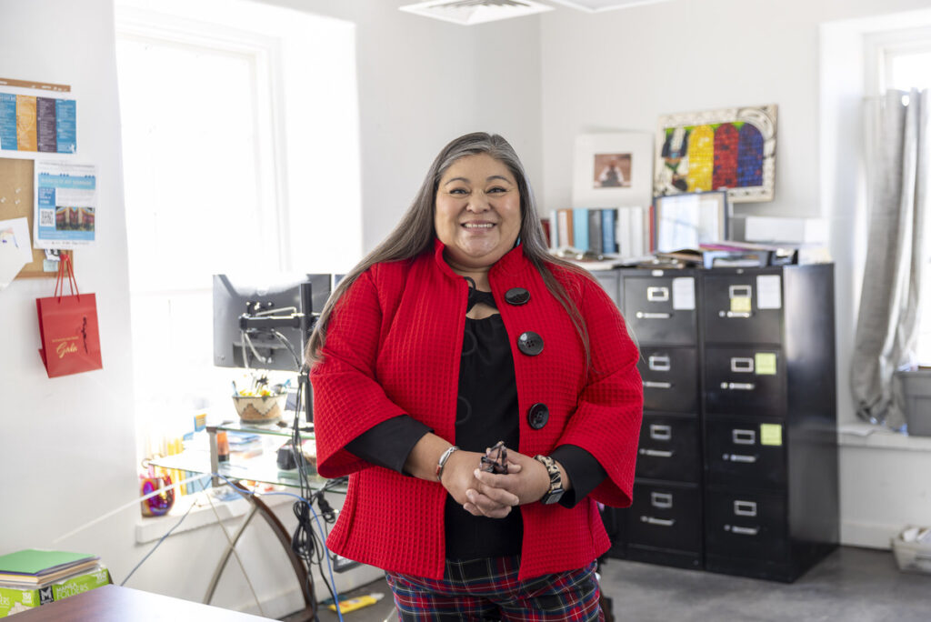 Mona Perea stands in an office setting. She is wearing a red textured jacket over a black top and plaid pants.