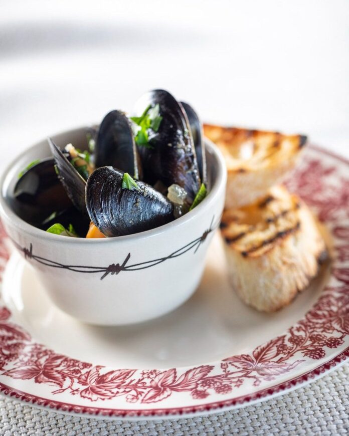 A small bowl hols Mussels in a broth that sit on a plate with two pieces of toasted baguette beside the bowl.