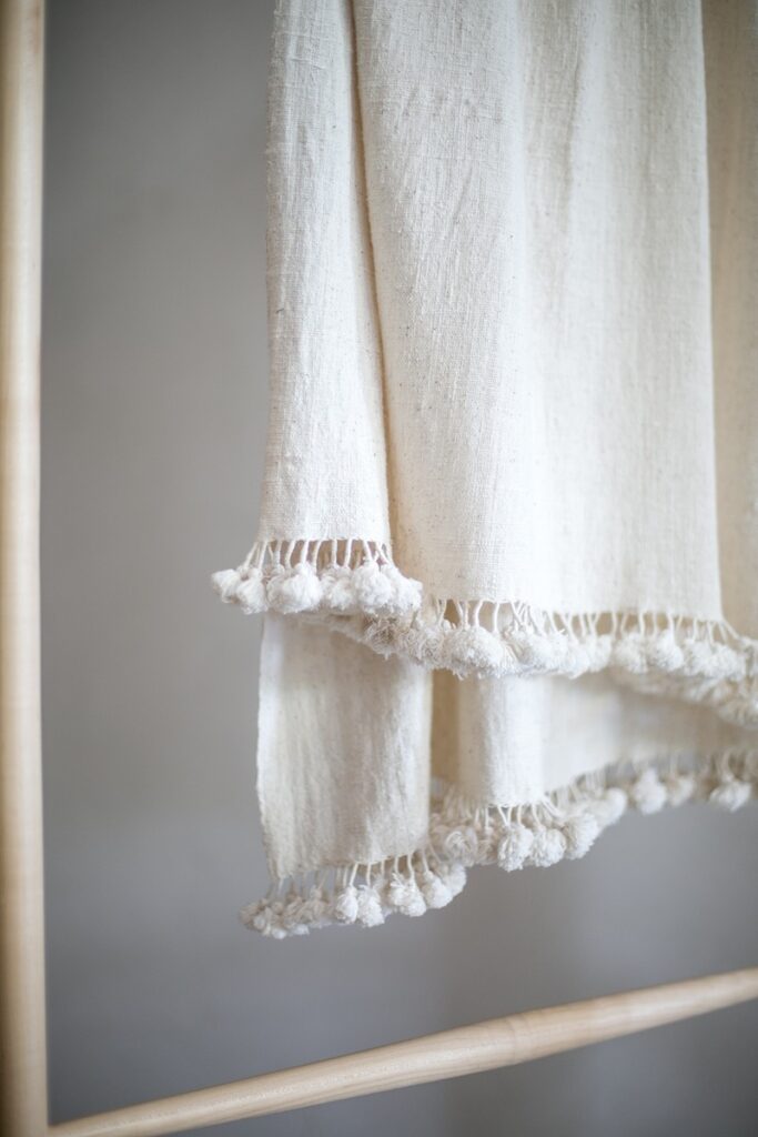 A close-up of a 100% undyed, organic Kala cotton throw or wrap with a textured surface and fringed edges with small pom-poms, draped over a wooden bar.