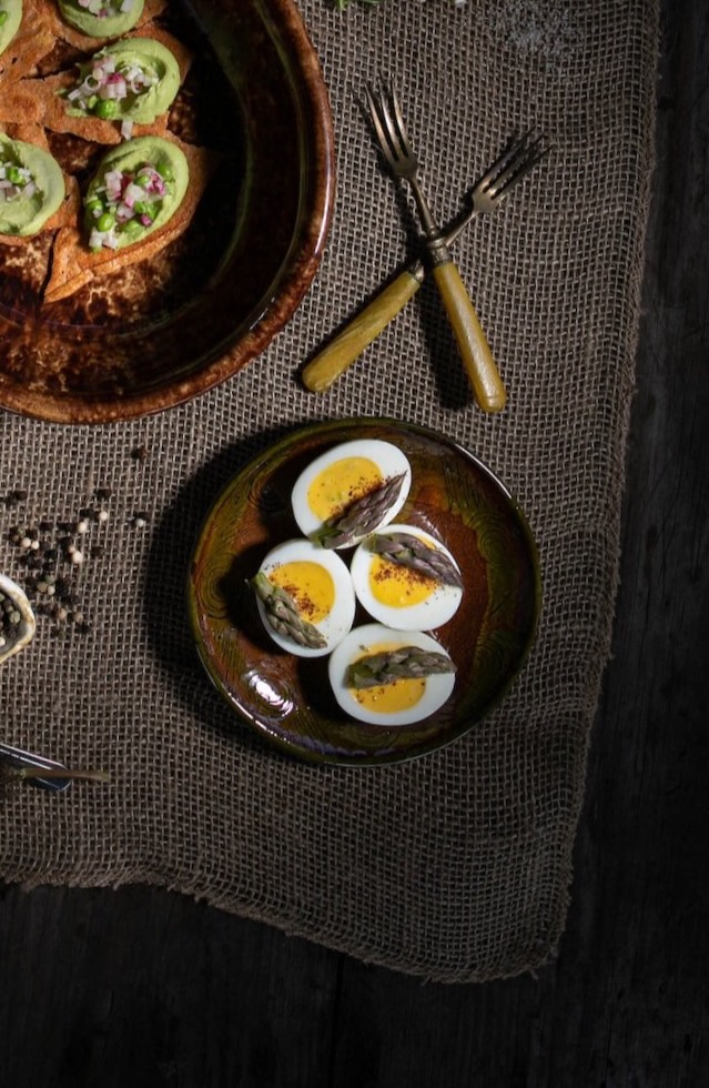 A small brown plate holds four deviled eggs topped with paprika as a plate of pea puree triangles sit in the upper left corner.