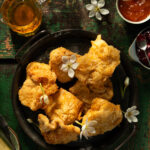 Paraoa Parai recipe shows golden brown pieces of fried bread, some adorned with small white flowers, arranged on a dark metal tray.