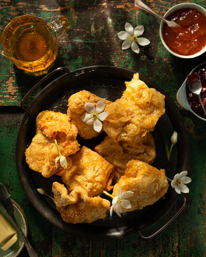 Paraoa Parai recipe shows golden brown pieces of fried bread, some adorned with small white flowers, arranged on a dark metal tray.
