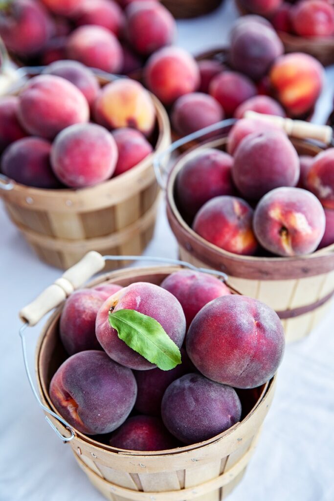 Wooden baskets filled with ripe peaches, some with a single green leaf, are displayed on a white surface.