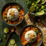 Two bowls of Ghanaian peanut soup with rice balls, garnished with chopped peanuts, okra, and cilantro, are presented on a green surface with lime wedges.