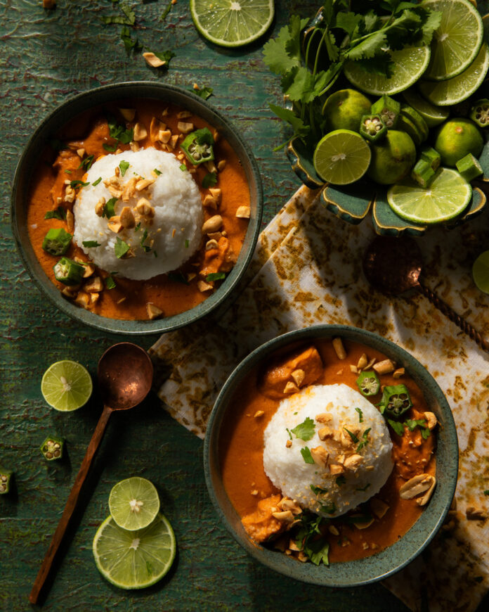 Two bowls of Ghanaian peanut soup with rice balls, garnished with chopped peanuts, okra, and cilantro, are presented on a green surface with lime wedges.
