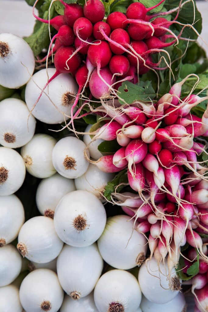 A close-up shot of bundles of red and white radishes with their leafy tops, alongside a pile of white onions.