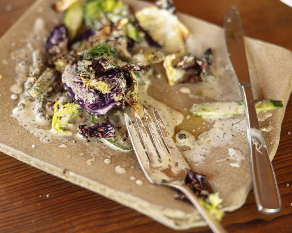 A fork and knife lay over a half eaten Roasted Cabbage Salad on a stone rectangular plate.