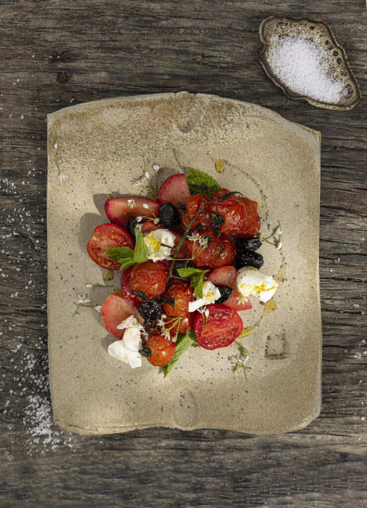 A close-up of a serving of roasted tomatoes still on the vine, accompanied by small balls of mozzarella and black olives.