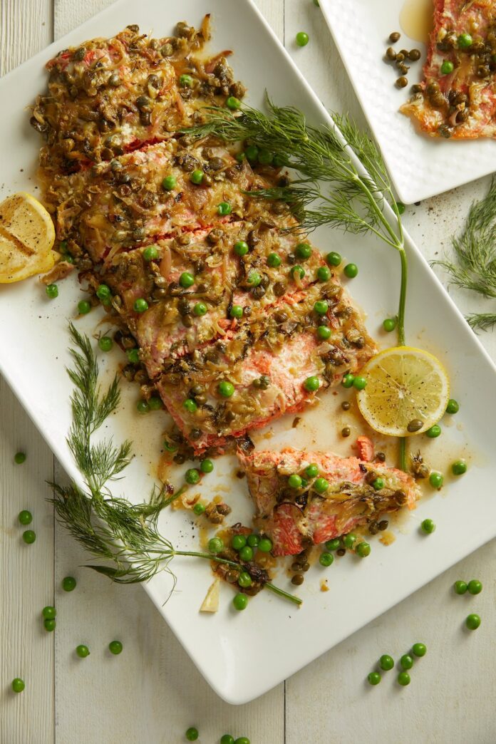 Salmon with Dill Butter Sauce and English Peas served on a white tray with herbs and lemons