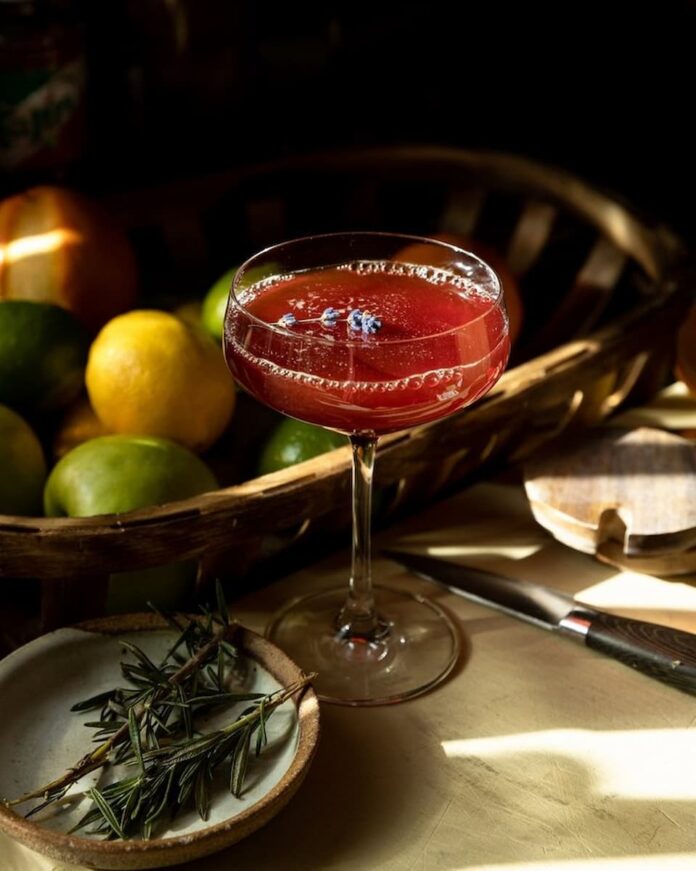 Sangre Sunset cocktail sits red in a coupe glass with a toothpick of blueberries floating on top.