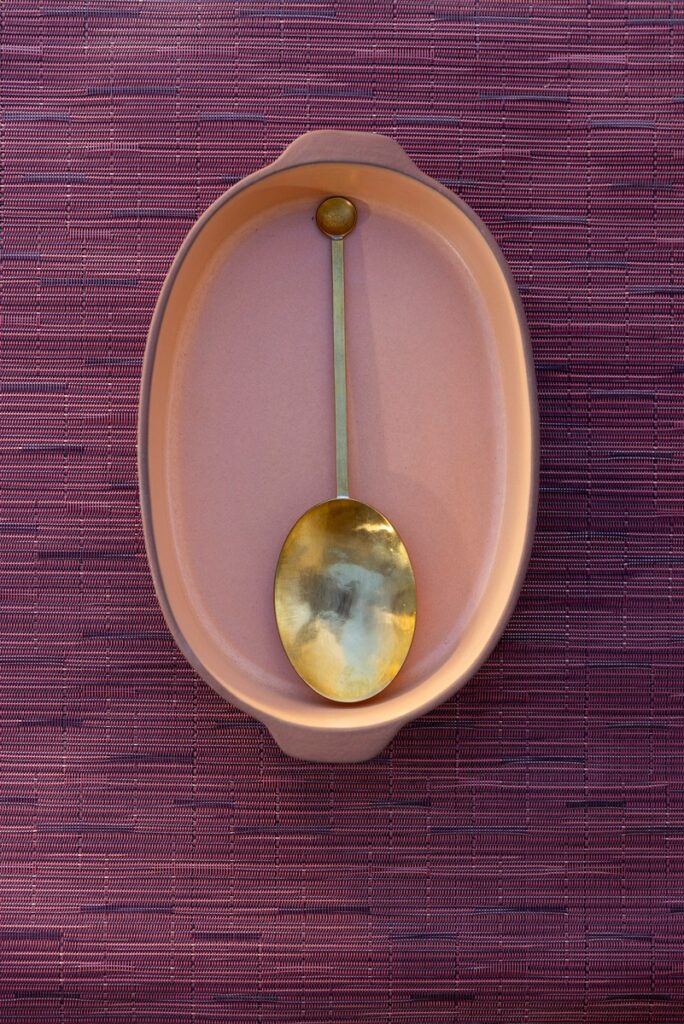 An overhead shot of a soft pink oval terracotta dish with a handmade brass spoon inside.