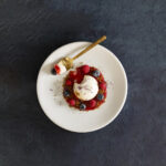 A beautifully plated dessert featuring a dome-shaped panna cotta, surrounded by a ring of graham crumble, fresh raspberries, and blueberries.