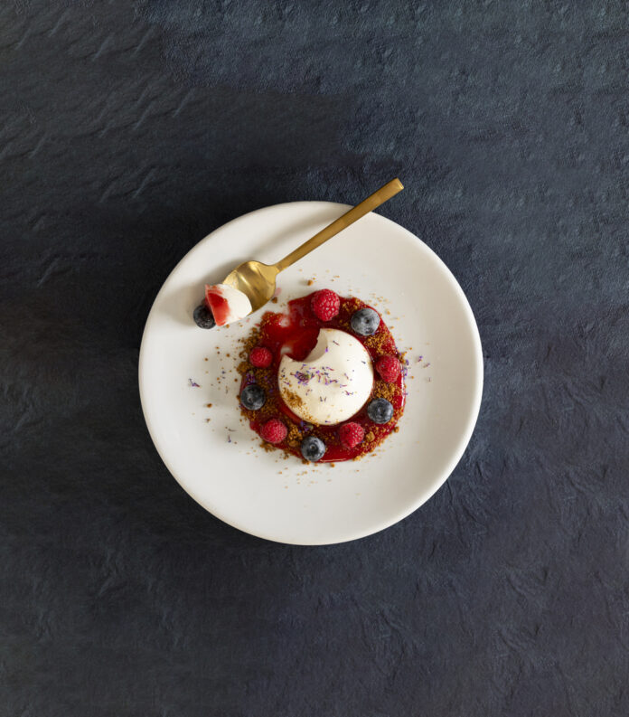 A beautifully plated dessert featuring a dome-shaped panna cotta, surrounded by a ring of graham crumble, fresh raspberries, and blueberries.