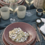 A red bowl with Spaghetti with Clams sits on a greenish tablecloth with a wine glass, candles, and another bowl of pasta behind it.
