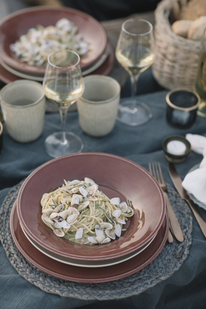 A red bowl with Spaghetti with Clams sits on a greenish tablecloth with a wine glass, candles, and another bowl of pasta behind it.