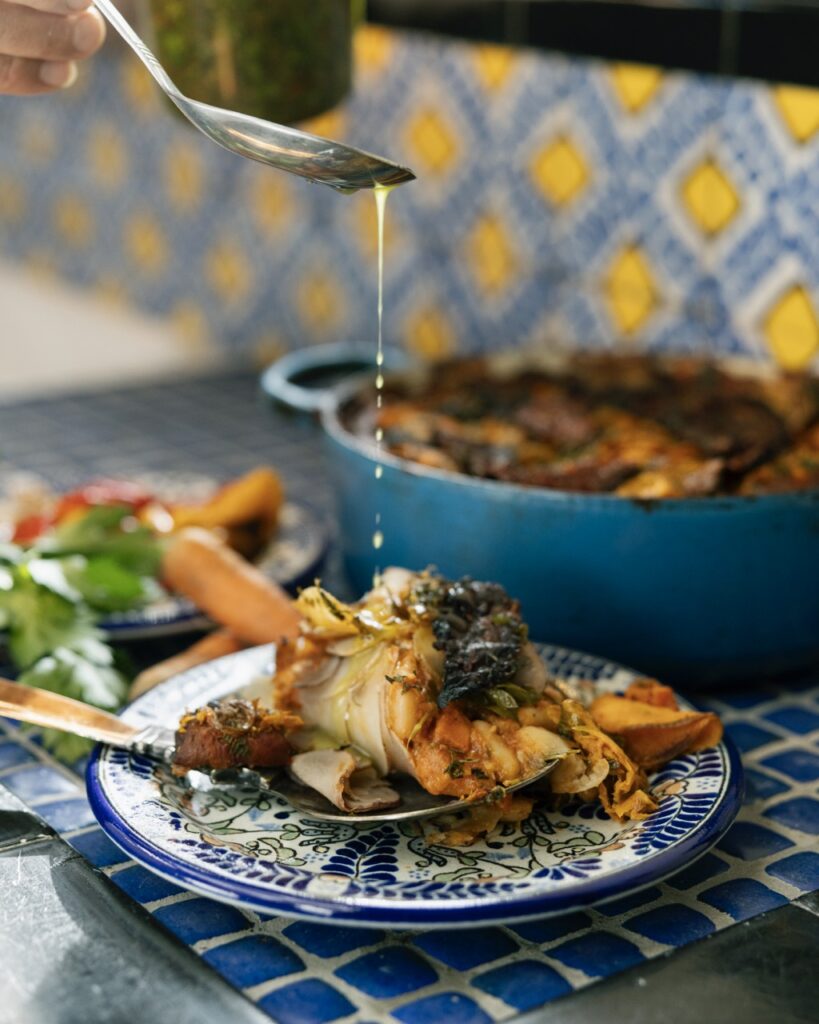 A blue plate holds a fish main dish as a person uses a spoon to drizzle a sauce on top.