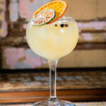 A Spanish Gin and Tonic cocktail, garnished with dried orange and lime slices and peppercorns, is presented in a large stemmed glass.