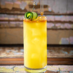 A vibrant sugar cane supper cocktail with a reddish-brown top layer is presented in a tall glass, garnished with a cucumber ribbon and a pipette.