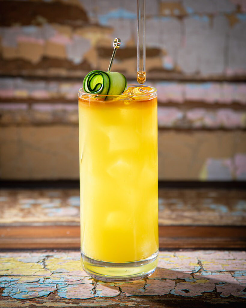 A vibrant sugar cane supper cocktail with a reddish-brown top layer is presented in a tall glass, garnished with a cucumber ribbon and a pipette.