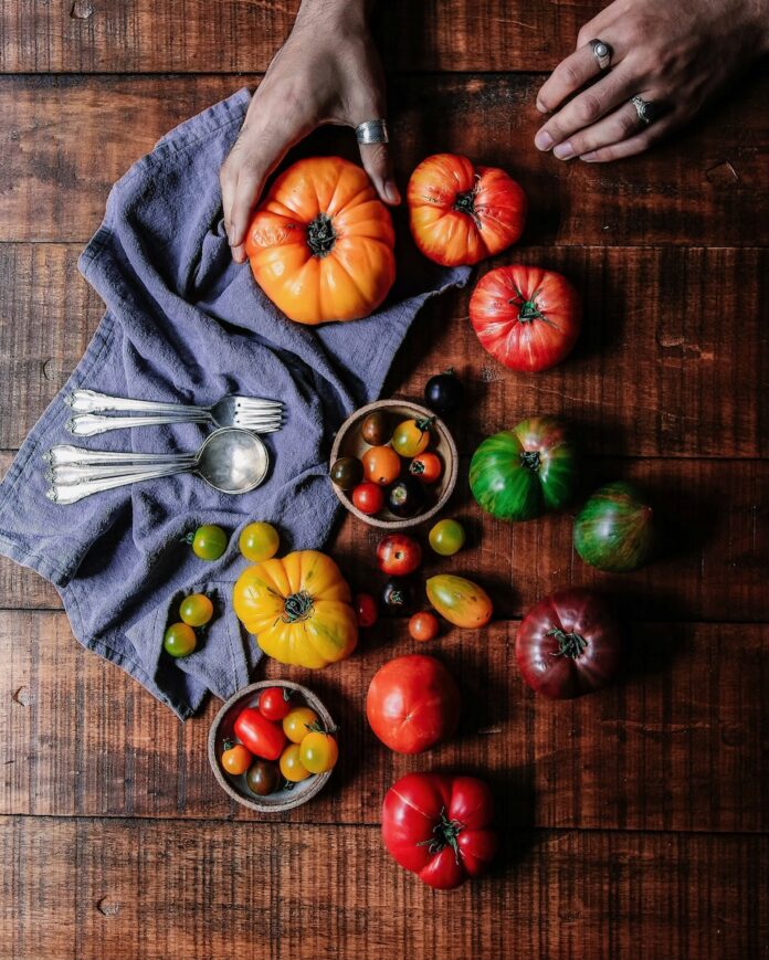 Various summer tomato varieties sit amongst a wood table with spoons to the left of them as a woman picks one up to make a recipe.