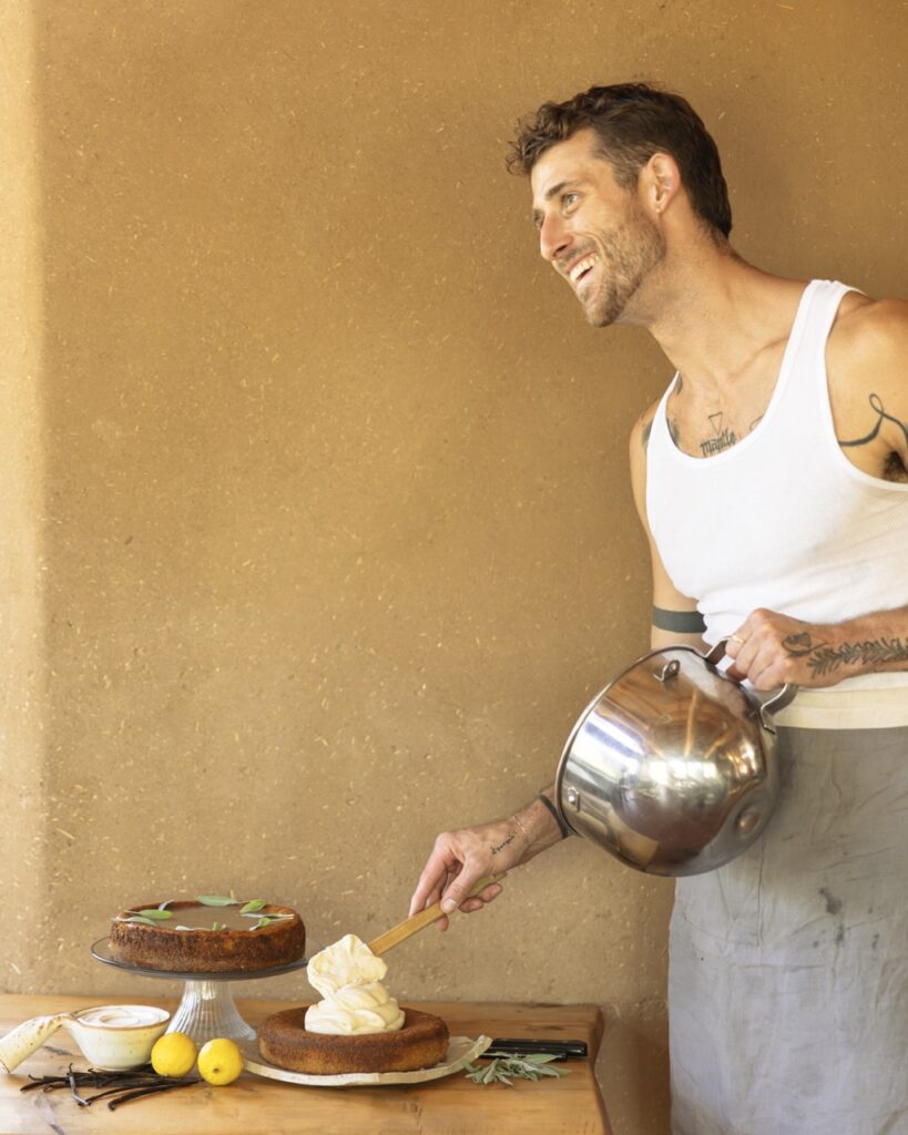 A man frosts a cake on a wood table against a neutral wall.